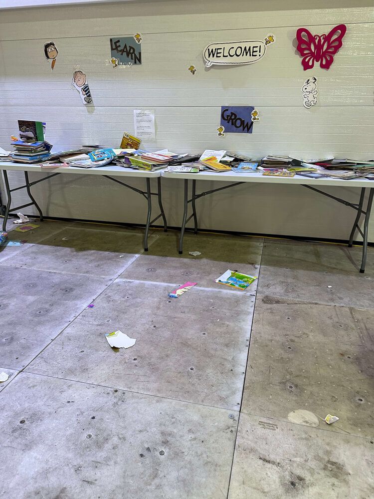 A photo taken at Floyd Bennett Field, an airport that has been converted into a shelter for new arrival families. It depicts a drab, concrete area with signs stuck on the wall that say “welcome”, “learn”, and “grow”, suggesting some sort of child enrichment or education space. There are two tables with a messy pile of old books in an otherwise very blank setting. Image credit: Coalition for the Homeless